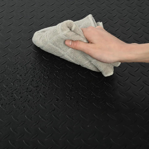 Close-up of black diamond plate design on Heavy Duty Anti Fatigue Floor Mat Built For Long Shifts 3 Ft By 7 Ft Thick Fifteen Sixteenths Inch Ergonomic Standing Mat For Commercial Kitchens And Workshop Floors being cleaned with a cloth.