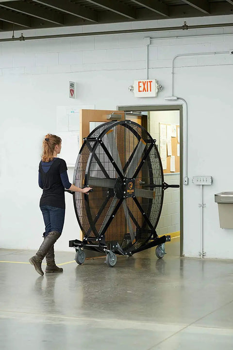 A person moving a 6 foot industrial mobile floor fan with OSHA certified safety cage wheels in a large workspace.