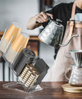 Clear Acrylic Cup And Lid Holder With Straw Organizer Adjustable Five Compartment Cup Dispenser For Disposable Paper And Plastic Cups The No Fuss Countertop Organizer For Your Home Kitchen with coffee brewing in the background.