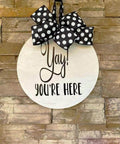 Round unfinished plywood sign with black script Yay! you're here, hung by a polka-dot ribbon.