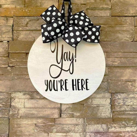 Round unfinished plywood sign with black script Yay! you're here, hung by a polka-dot ribbon.