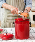 Person using red metal pail for countertop waste recycling from Outdoor Dual Chamber 43 Gallon Compost Tumbler Kit With 1.5 Gallon Kitchen Bin Lid And Charcoal Filter Rotating Design For Easy Separation Black And Red Metal Pail For Countertop And Outdoor Waste Recycling.