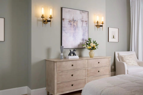 Two-Light Gold Brass Wall Sconces with 2.8 Inch Cylindrical Clear Glass Shades, E26 Base, in a bedroom setting on pale wall.