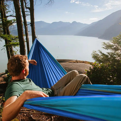 Person relaxing in Neolite Single Camping Hammock Ultra Lightweight Portable Nylon Parachute Hammock by lakeside, sky blue color.
