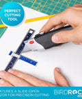 Retractable utility knife with black rubber handle cutting a blue ruler on white surface; BirdRock Home.