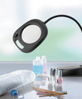 Go-To LED Floor Lamp And Table Lamp With 5-Inch Magnifier on a salon table with beauty products, flexible arm visible, sleek black design.
