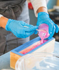 Person pouring pink soap mixture into Ultimate 7 Piece Soap Making Kit With Silicone Molds And Stainless Steel Slicers, wood box visible