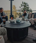 People gathered around a Surround Surface Fire Pit Lid For Bonfire And Ranger Pits Built For The Backyard Boss Vibe Durable Cold Rolled Steel Cover 23.75 Inch Diameter 7.05 Lb Weight Converts To A Sleek Outdoor Tabletop on a deck.