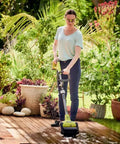 Woman using Cordless 18 Volt Patio Surface Cleaner With Telescoping Handle on a wooden deck surrounded by greenery.
