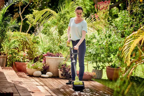 Woman using Cordless 18 Volt Patio Surface Cleaner With Telescoping Handle on a wooden deck surrounded by greenery.