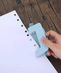 Hand using a two hole paper punch metal blue desktop puncher for file folders and loose leaf books, showing punching action on white paper.