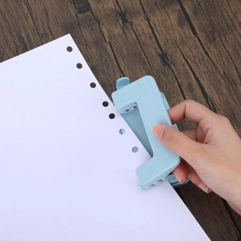 Hand using a two hole paper punch metal blue desktop puncher for file folders and loose leaf books, showing punching action on white paper.