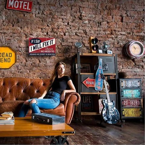 Vintage Metal Man Cave Sign For Men If I Say I Will Fix It There Is No Need To Remind Me Every 6 Months Funny Garage Wall Decor For Workshop Basement And Home Bar 12 X 8 Inches displayed in a stylish room with brick walls, next to a leather couch and guitar.