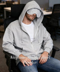 Man wearing Unisex Travel Hoodie With Built In Eye Mask In Airplane Mode For Naps On The Fly Cozy Zip Up Fleece Jacket in grey sitting.