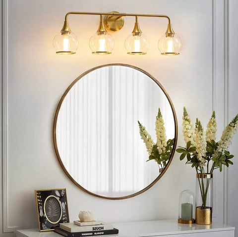 Hot Gold 4 Light Vanity Light Fixture Over Mirror With Clear Glass Globe Shades 30.7 Inch Wall Sconce Bath Lighting For Master Bathroom Bulbs Not Included. Gold fixture above round mirror and flowers on white wall.
