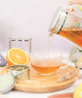 Top Tier Double Walled Insulated Teapot That Keeps Your Daily Brew Sizzling Hot With Slick Stainless Steel Filter Lid For Loose Leaf Tea pouring tea into a glass cup surrounded by citrus fruits.