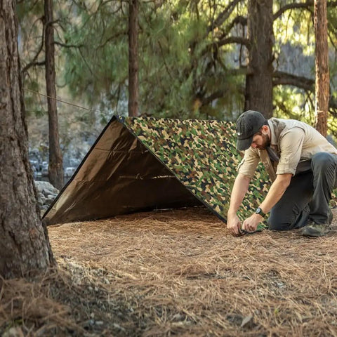 Man setting up Rugged Survival Tarp With Insulated Reflective Mylar Layer For All Weather Camping, woodland camo tarp in forest