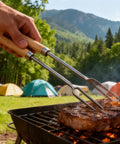 Person using stainless steel BBQ forks for grilling steak at a campsite with tents in background, showcasing heat resistant handle.