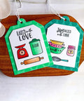 Two decorative kitchen-themed tags with baking illustrations and phrases 'Baked with Love' and 'Jam Packed with Love' on a wooden board.