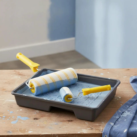 Knockout Paint Roller One Pack With A Legit Synthetic Fiber Cover And Dual Yellow Stripes To Slam Missed Spots And Serve Up Butter Smooth Coverage For Any Weekend Warrior DIY Project in a paint tray with blue paint.