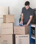 Person using Six Roll Pack Of High Performance Clear Packing Tape Refills For Shipping Moving Mailing And Storage Boxes, 1.88 Inch Wide By 60 Yard Length, 3 Inch Core, All Weather Grip And Super Strong Adhesive to seal boxes in a garage setting.