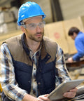 Man in workshop wearing Unisex Safety Glasses Bulk Pack 84 Pairs With Polycarbonate Lens, holding tablet, wearing blue hard hat