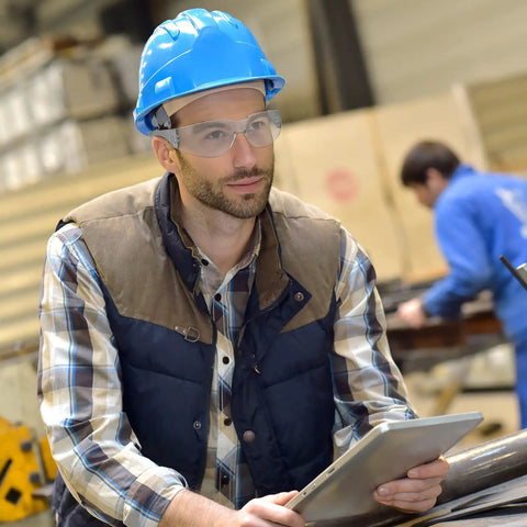 Man in workshop wearing Unisex Safety Glasses Bulk Pack 84 Pairs With Polycarbonate Lens, holding tablet, wearing blue hard hat