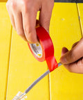 Hands applying red PVC Electrical Insulating Tape Roll Built To Last With Strong Adhesion And Abrasion Resistance For Industrial Equipment Maintenance Antiaging Shield For Safe Electrical Insulation In Harsh Environments around a grey cable on a yellow table surface.