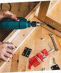 Person using a green power drill on a wooden frame with various hardware and tools including hinges and a red clamp.