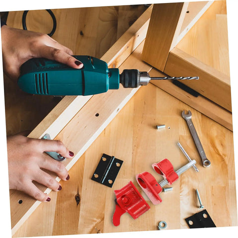 Person using a green power drill on a wooden frame with various hardware and tools including hinges and a red clamp.
