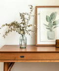 Wooden table with a vase of dried branches, framed plant artwork, and candles