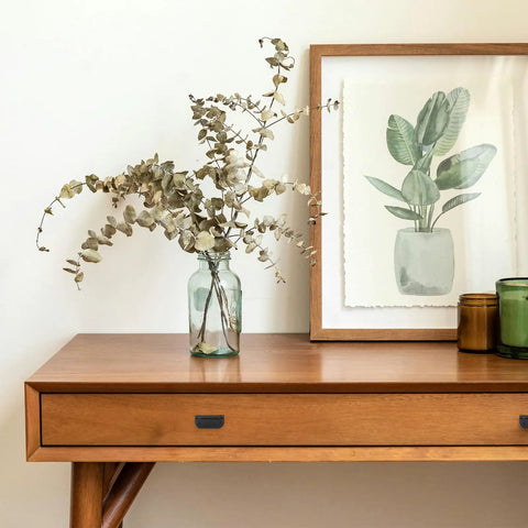 Wooden table with a vase of dried branches, framed plant artwork, and candles