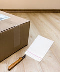 Fifty Pack Small Bubble Mailers 10x10 Plus 3cm Self Seal Padded Envelopes next to cardboard box and knife on wooden floor