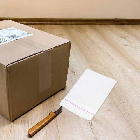 Fifty Pack Small Bubble Mailers 10x10 Plus 3cm Self Seal Padded Envelopes next to cardboard box and knife on wooden floor