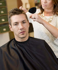 Front view of man wearing a black barber cape as stylist uses neck duster brush and barber brush