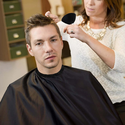 Front view of man wearing a black barber cape as stylist uses neck duster brush and barber brush
