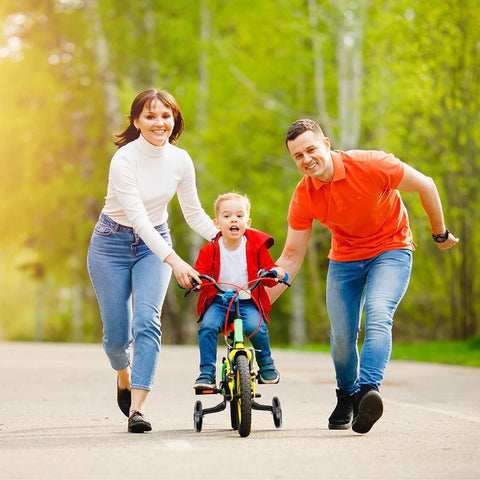 Child riding a bike with Quiet Training Wheels Kit for 12-20 inch kids bikes, guided by parents on a sunny day.