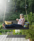 Woman relaxing on an upgraded patio wooden porch swing for courtyard and garden, heavy duty 880 lbs swing chair with black finish.