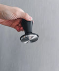 Hand holding Spring Loaded Espresso Tamper 53.3mm with ripple base and anodized aluminum handle, side view.