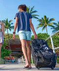 Person carrying a Roller Mesh Dive Gear Bag With Large Outer Pocket And Internal Pocket, Adjustable Shoulder Strap, Heavy Duty Travel Roller Bag for Scuba Gear, 29.9x13.8x11.8 Inches Black, on a tropical boardwalk.