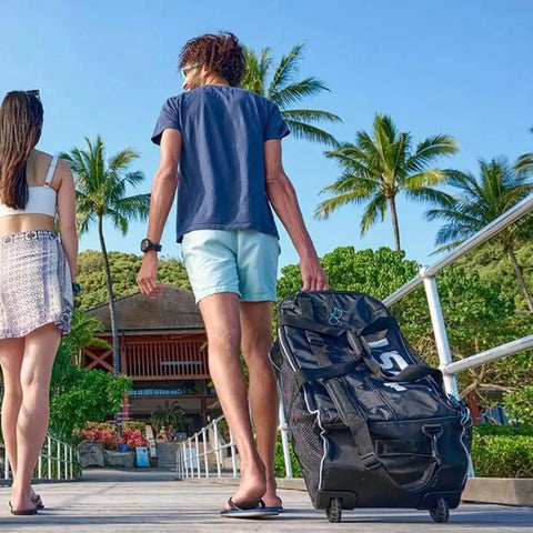 Person carrying a Roller Mesh Dive Gear Bag With Large Outer Pocket And Internal Pocket, Adjustable Shoulder Strap, Heavy Duty Travel Roller Bag for Scuba Gear, 29.9x13.8x11.8 Inches Black, on a tropical boardwalk.