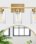 Clean Three Light Bathroom Vanity Wall Sconce In Smooth Brushed Gold Finish With Clear Retro Glass Shades, viewed from front with mirror.