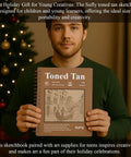 Man holding a Tan Toned Sketchbook For Mixed Media With Sixty Sheets Of 90lb 150gsm Tan Drawing Paper 5.5 By 8.5 Inches Great For Charcoal Graphite Colored Pencils And White Gel Pens One Hundred Percent Recycled Wire Bound in front of a decorated Christmas tree.