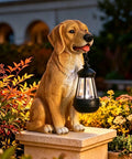 Golden Retriever Solar Dog Statue For Outdoor Decor With Lantern Lights Backyard Lawn Yard Art Weatherproof Garden Ornament For Patio Sun Powered Lights Lit Housewarming Gift on pedestal among flowers.