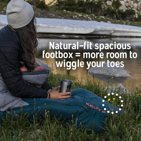 Person sitting in a Cosmic Synthetic Fill 40 Degree Backpacking Sleeping Bag With Compression Straps in nature scene.