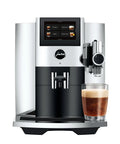 Premium Fully Automatic Chrome Espresso Coffee Machine Featuring A Hella Smooth Four Point Three Inch Smart Touchscreen Display For Twenty Seven Freshly Ground Drinks, black and chrome with a glass of espresso.