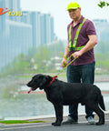 Man wearing reflective gear and neon yellow cap walking black dog on urban path