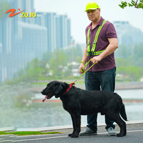 Man wearing High Visibility Reflective Running Vest Pack of 10 Neon Yellow outdoors walking a black dog with cityscape backdrop.
