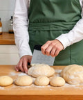 Front view of a 6-inch stainless steel dough scraper with a black handle and measuring scale, cutting dough on a wooden countertop.