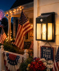 Outdoor Solar Porch Lights 2 Pack Featuring Motion Sensor And 3 Lighting Modes Zero Wiring Hassle Built Tough Weatherproof Wall Lanterns Sconces For Front Door Garage Fence Patio 7 5 Inch mounted beside flowers and American flags on porch at night.